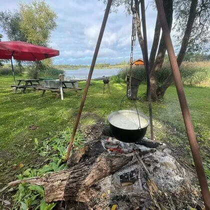 Katls karājas virs ugunskura, fons ar Daugavu un sēdvietām.