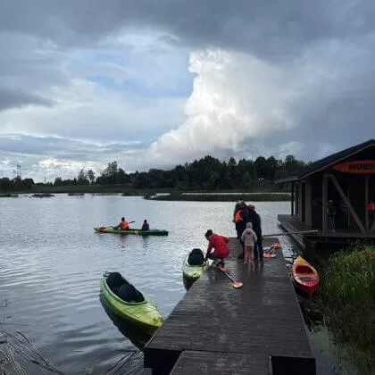 Cilvēki ar kajakiem pie piestātnes mākoņainā dienā.